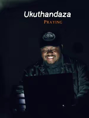 The image shows a man in a dark setting, likely indoors, working on a laptop. He's wearing a black cap and a jacket. The backdrop is dim with a gradient of shades, making the man's face stand out due to the screen's glow, highlighting his focused expression.. Above him is text which reads, "Ukuthandaza" with the word "Praying" below. The background is dark.