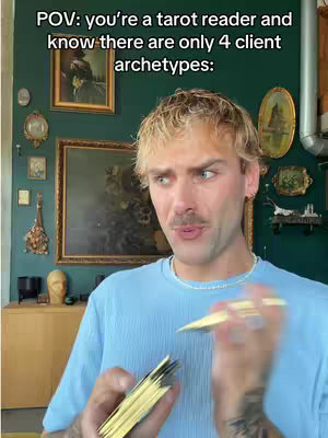 A man with light hair and a mustache is making a face while holding a deck of tarot cards. The background is a dark green wall with various framed paintings and decorative objects. The man is wearing a light blue shirt, and the image has a slightly desaturated look.