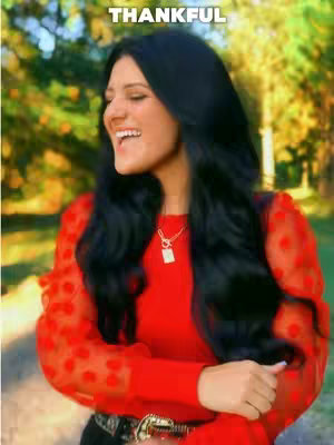A woman with long black hair smiles, wearing a red top. The background features blurred greenery. Text overlayed at the top reads 'THANKFUL'.
