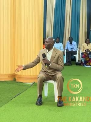 A man in a suit sits on a chair addressing a group. He holds a microphone, gesturing with his right hand outstretched. The background features a yellow wall on the left, and a blue-and-white curtain on the right. The floor is green, with a logo in the bottom right.