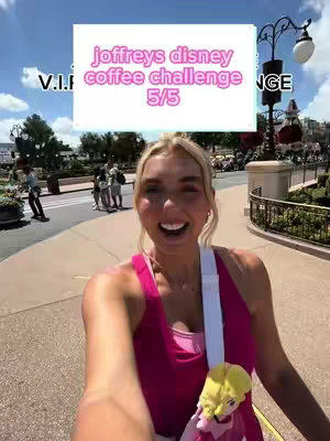A woman with blonde hair smiles at the camera while walking down a path. She wears a pink tank top and has a white strap across her chest. There is a white box overlay in the upper center of the image with pink text. In the background, there are trees, a street, and other people. She is also holding a small plush figure.