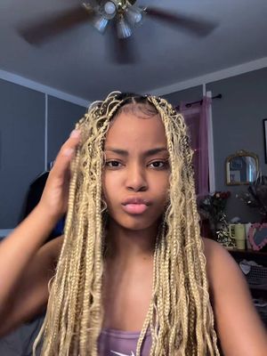 A selfie-style shot featuring a young woman with long, blonde braided hair. She is looking directly at the camera with a slight pout. Her expression is neutral. She is in a room; the ceiling fan is visible.