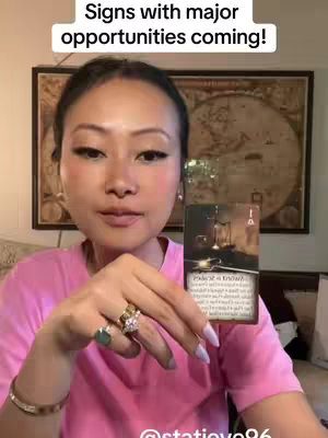 A woman with fair skin and dark hair styled away from her face is centered in the frame. She wears a pink t-shirt, multiple rings on her left hand. She is holding a tarot card in her right hands, gazing forward. The background shows a framed artwork and a glimpse of a wall. Overhead text identifies the content related to astrology.