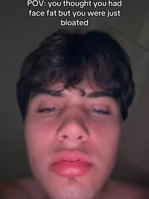 A close-up shot of a young man's face, with a focus on his puffy, full lips and slightly closed, tired-looking eyes. Text overlaid at the top reads, 'POV: you thought you had face fat but you were just bloated.' The lighting is dim, and the composition is centered on the man's face.