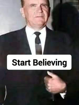 A man wearing a dark suit and tie is the central figure in the image. The text overlayed at the bottom reads, 'Start Believing.' The background is slightly blurred but appears to be a hallway.