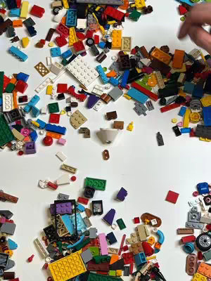 A top-down shot of various colored Lego bricks scattered on a white surface. There are numerous bricks of all shapes and sizes, with a hand partially visible at the top right corner. The composition is busy and colorful.
