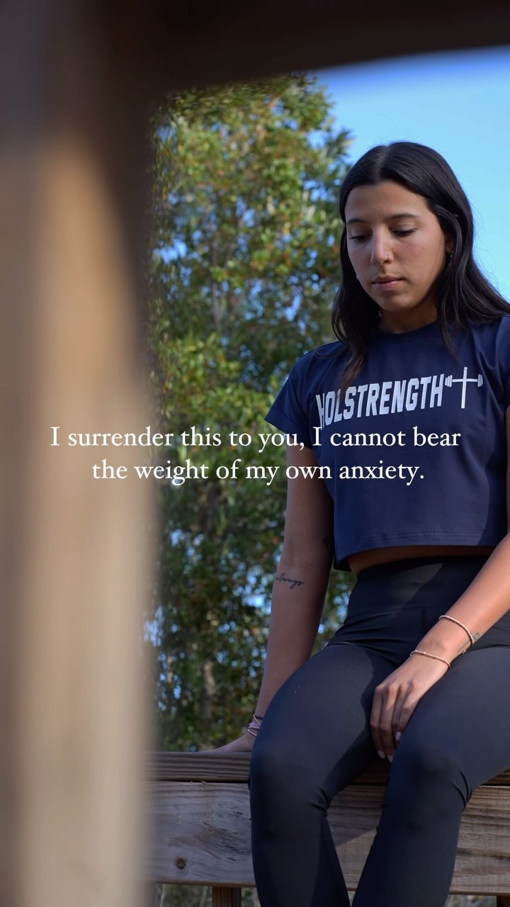 A woman is shown in a serene outdoor setting, seated on a wooden structure with trees and sky in the background. She wears a navy crop top and black leggings and has a focused expression.