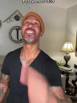 A man with a gray beard and an orange beanie is smiling up at the camera. The image is lit by warm lighting, and the background shows a decorated wall with a large letter "T" over a fireplace mantel. The man is wearing a black shirt, and a hand is visible at the bottom of the frame.