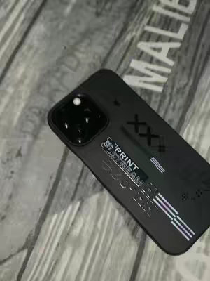 A close-up shot of an iPhone with a black case, angled against a wooden background with the word 'MALIBU' visible. The phone case has graphic text and horizontal lines. The overall composition is angled to the right.