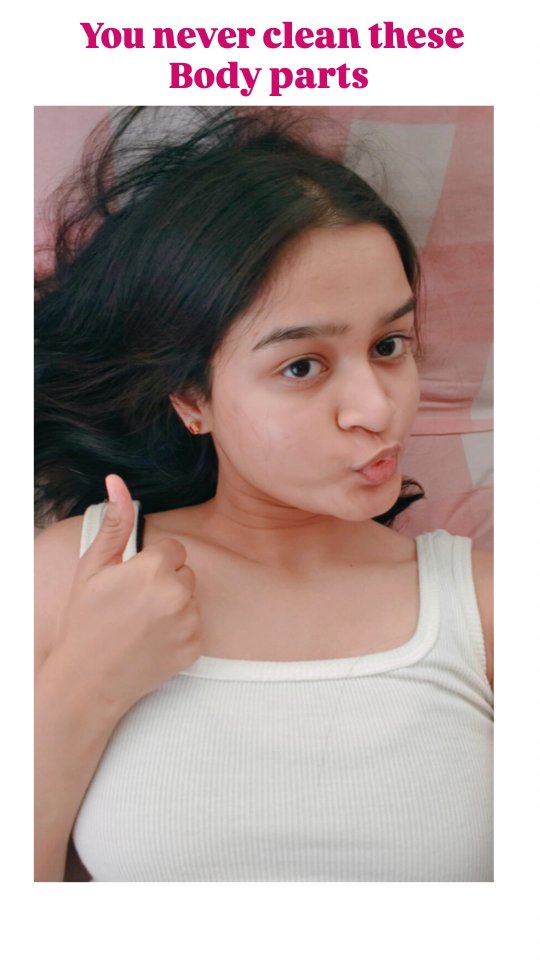 A selfie of a young woman lying on a bed. She has dark hair and is wearing a white tank top. She is making a kissing face with her lips and giving a thumbs up. The background is a light pink color.