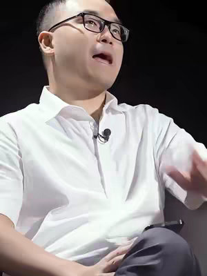 The image shows a man in a white dress shirt and glasses, speaking directly toward the camera, with a black background. A microphone is clipped to his shirt.