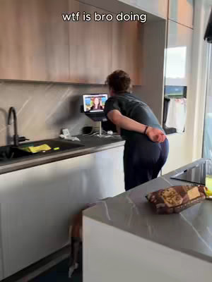 A person stands in a modern kitchen, their back to the camera, seemingly observing something on a laptop perched on a stand. The kitchen features light-colored cabinets, a dark sink, and a contrasting light countertop. A small dog is visible near the person.