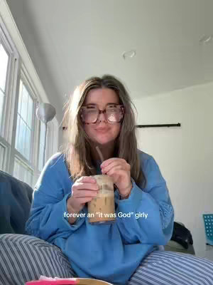 A woman with light brown layered hair and large glasses sits indoors with a drink. The woman is smiling while holding a clear glass with a straw. The lighting is bright, and a window is in the background.