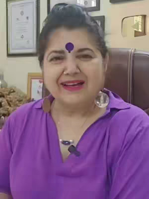 A woman with dark hair, wearing a purple top and earrings, is looking at the camera with a smile. She has a bindi on her forehead. The background shows an office setting with framed items.