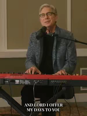 The cover image features a man playing a red-keyed keyboard in front of a microphone. Behind him is a blurred background with a window and a light-colored wall. Text overlays the bottom, stating 'AND LORD I OFFER MY DAYS TO YOU'. The man is wearing glasses and a grey jacket, appearing to be in a performance setting.
