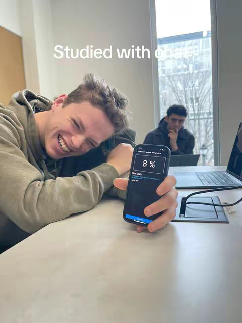 A young man in a casual hoodie smiles broadly, holding a smartphone that displays an app screen, with a light-colored table and another person in the background near a window.