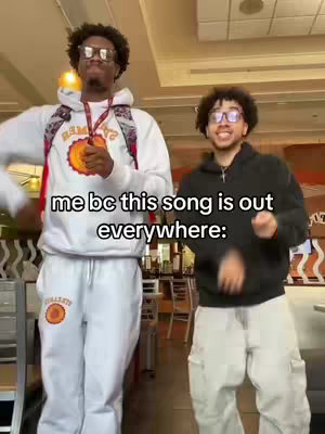 Two young Black men are captured mid-dance in a casual indoor setting. They are both wearing glasses and hoodies, with a text overlay on the image. The man on the left is wearing a white hoodie and sweatpants with some form of logo on it, and the man on the right is wearing a black hoodie and gray cargo pants. The background appears to be an eatery setting with ceiling lights.