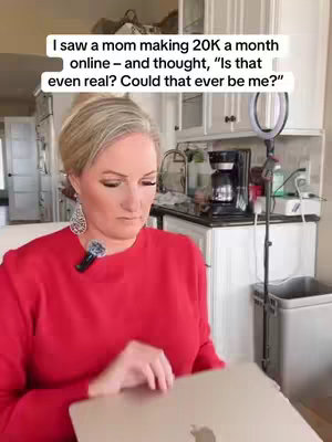A woman in a red long-sleeved top is seated indoors, holding a silver laptop. Her short, blonde hair is neatly styled. She wears drop earrings and has a microphone clipped to her collar. The background shows a kitchen setup, with white cabinets and appliances. Overhead, there's a ring light.