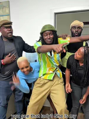 A group of five people are in a room. The center person is pointing, with arms around others who are reacting with different expressions. Two other people are also gesturing with their arms.