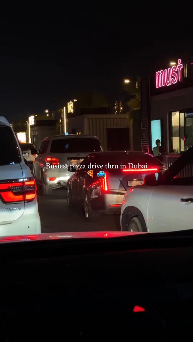 The cover image is taken from inside a car, showing a long line of cars waiting, likely at a drive-through. The scene unfolds at night, illuminated by the headlights and the signage of a food establishment called "must".  The text overlay on the image indicates the image is a busy drive-thru at this pizza place located in Dubai.