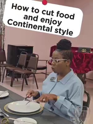 A woman in a light blue shirt sits at a table, demonstrating how to cut food. She is holding a knife and fork over a plate set on a table. Text overlayed states 'How to cut food and enjoy Continental style'. The background includes chairs, a table with a red cloth, and a dark area that could be a cabinet or a speaker.