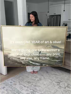 A woman is holding a large framed artwork. The artwork is a landscape painting. Above the artwork, in white text, is the video's hook. The woman is standing on a patterned rug, and the background is a modern living room with white walls.