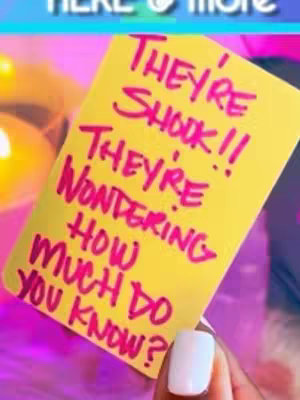 A hand holding a yellow sticky note with handwritten text in pink marker. The background is blurred with shades of pink and purple.