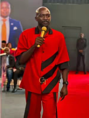 A man in a red suit with black stripes is holding a golden microphone, speaking on a stage. There is a blurred background of people and a projection screen.