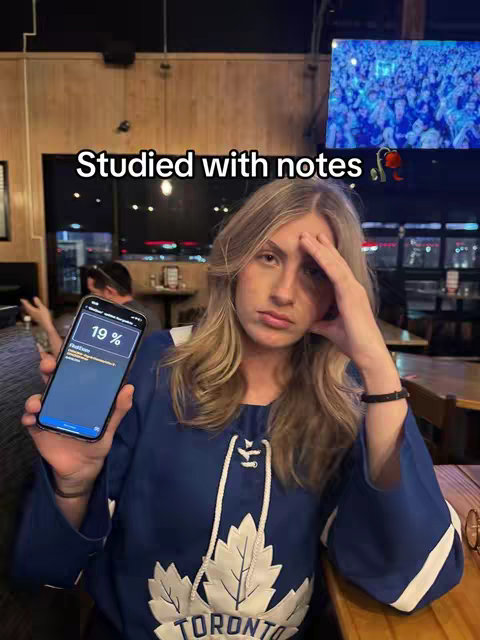 A selfie-style shot of a person in a sports jersey, inside a bar or restaurant setting. The person appears to be looking at the camera with a disappointed expression, and holding a smartphone displaying a low percentage on the screen. The background includes tables, chairs, and a large screen possibly showing a crowd.