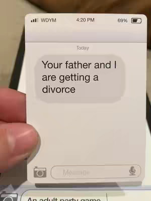 The image is a close-up of a white card styled to look like a phone text message. The card shows a mock iPhone interface. In the center of the card, a large text bubble displays the message: "Your father and I are getting a divorce". The top of the card mimics a phone's status bar, displaying the carrier name, time (4:20 PM), and battery percentage. Below the message, there's a simulated text input field with a camera icon on the left and the word "Message" displayed. The bottom of the card is partially visible with the words "An adult party game". A hand is partly visible at the bottom left corner, holding the card.