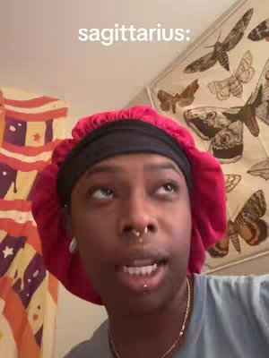 A close-up shot of a person, wearing a red cap, black headband and a blue top with a necklace in the background is a decorated wall. A text overlay that says "sagittarius:"