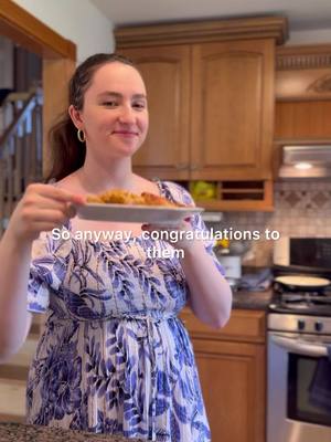 A woman in a floral dress holds a plate of food in a kitchen, smiling at the camera. Text overlayed says, 'So anyway, congratulations to them.' The setting includes wooden cabinets and stainless steel appliances.