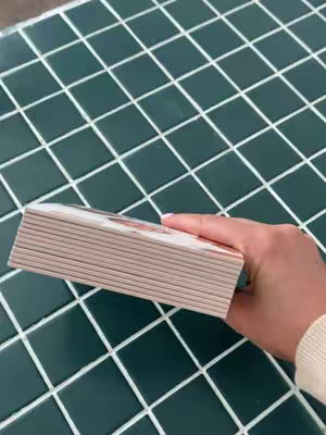 A hand holding a stack of small, rectangular photo journals. The hand and journals are positioned over a dark green, tiled surface. The journals are light-colored, and the background has a grid-like pattern.