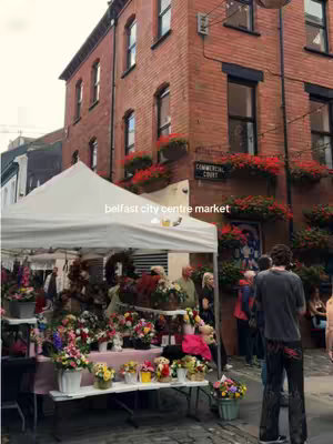 A bustling outdoor market scene in Belfast, featuring a white tent with various colorful flower arrangements on display in front of a brick building. People are milling about, and the scene has a vibrant, lively atmosphere.
