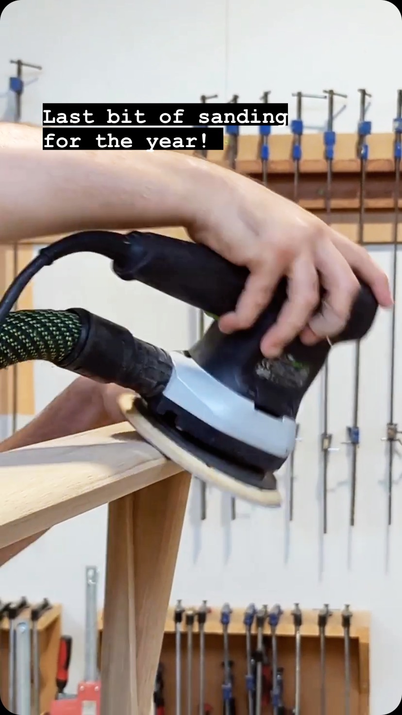 A person is sanding a piece of light-colored wood with a power sander. The sander is black and gray; the person's hands operate it. In the background, a wall has multiple clamps hanging. Text overlayed on the image has the phrase 'Last bit of sanding for the year!.'