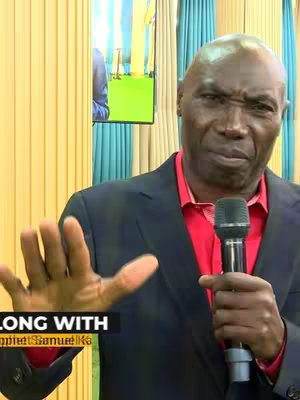 A man in a suit and red shirt is holding a microphone. He is gesturing with his left hand as if making a point. There is a screen behind him with a green and yellow background.