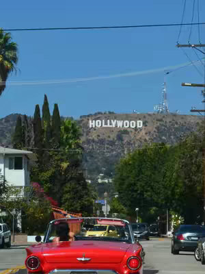 A photo taken from inside a red convertible car, driving down a city street. In the background, the HOLLYWOOD sign is visible on the side of a hill under a clear blue sky. Other cars are driving in the same direction on the street.