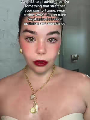 A close-up shot of a fair-skinned woman with dark hair and a slight smile. She has pronounced blush on her cheeks and deep red lipstick. She wears gold earrings and a gold necklace with a heart and medallion pendant. Text overlayed on top reads: "Say YES to all adventures. Do something that stretches your comfort zone. Wear citrine. why? Jupiter ruled Sagittarius thrives on optimism and abundance".