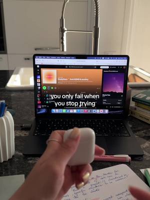 A laptop displaying a music playlist on a countertop, with a hand holding earbuds in the foreground. The background includes a kitchen faucet and a notebook with handwritten notes. The image has a warm, natural lighting.