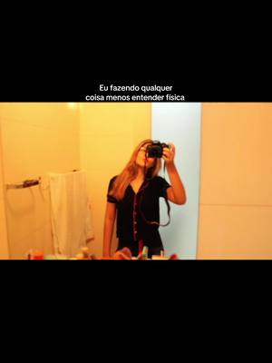 A young woman in a bathroom takes a selfie with a camera, viewed through a mirror. She has long blond hair and is dressed in black. Cosmetics and toiletries are visible on the counter.