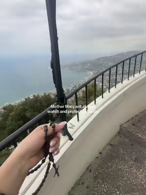 A scenic view from a high vantage point overlooking a coastal city and the sea. In the foreground, a hand holding a rosary is visible. The sky is overcast, and the image has a slightly muted color palette.