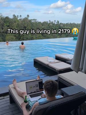 A man is seen lounging on a chair next to an infinity pool, working on a laptop while holding a beer. In the background, other people are swimming and the view extends into an endless natural landscape.