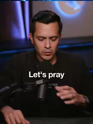 A man with short dark hair looks slightly down, illuminated by soft light. He wears a dark blazer and is positioned in front of a microphone. Text 'Let's pray' is superimposed over his chest in white font.