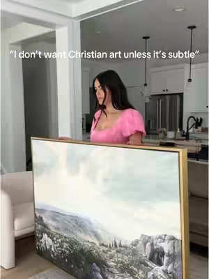 A woman in a pink dress holds up a large painting. She is standing in a modern, minimal living space with white walls and a light-colored floor.