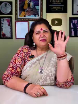 A woman with a red bindi in the center of her forehead is sitting on a chair and making a hand gesture. She is wearing a pale white dress with a floral design on a red top. Behind the person are awards and pictures. The hand is raised with her fingers spread.