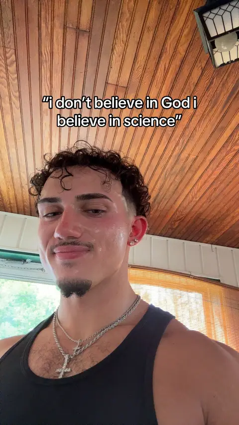 A close-up shot features a young man looking at the camera with a slight smile. He has short, curly hair and a trimmed beard. He's wearing a black tank top and a silver chain with a cross pendant. The background shows a wood-paneled ceiling.