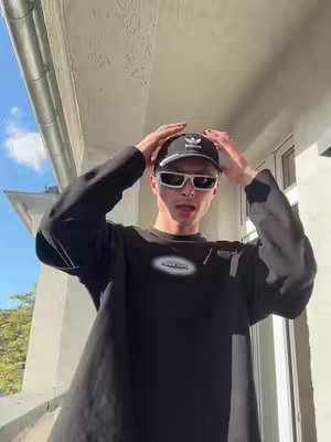 A young man wearing a black outfit, a black cap with a white logo, and white sunglasses stands on a balcony. He has his hands up, touching the cap. The composition is centered, and the natural light suggests a sunny day.