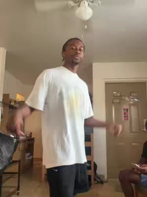 A man in a white t-shirt and black shorts is standing in a room with his arms to the side. Behind him is a door and a ceiling fan.
