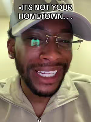 A close-up shot of a Black man smiling at the camera. He's wearing a light gray hat and glasses. The overlayed text says, '.ITS NOT YOUR HOMETOWN...' He has a short beard and is wearing a beige zip-up jacket.