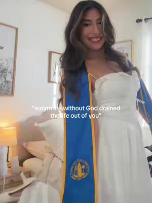 A young woman, likely a college graduate, stands indoors smiling. She is wearing a white dress and a blue and gold graduation sash. Behind her is a room with cream-colored walls, framed artwork, and a light fixture. The overall tone is celebratory and positive with a focus on the woman's happiness.